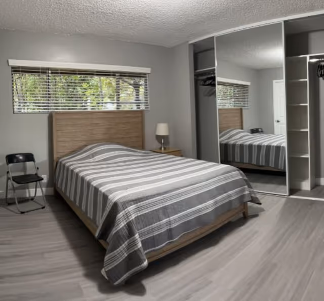 A furnished bedroom with a bed, nightstand, lamp, window blinds, and mirrored closet.