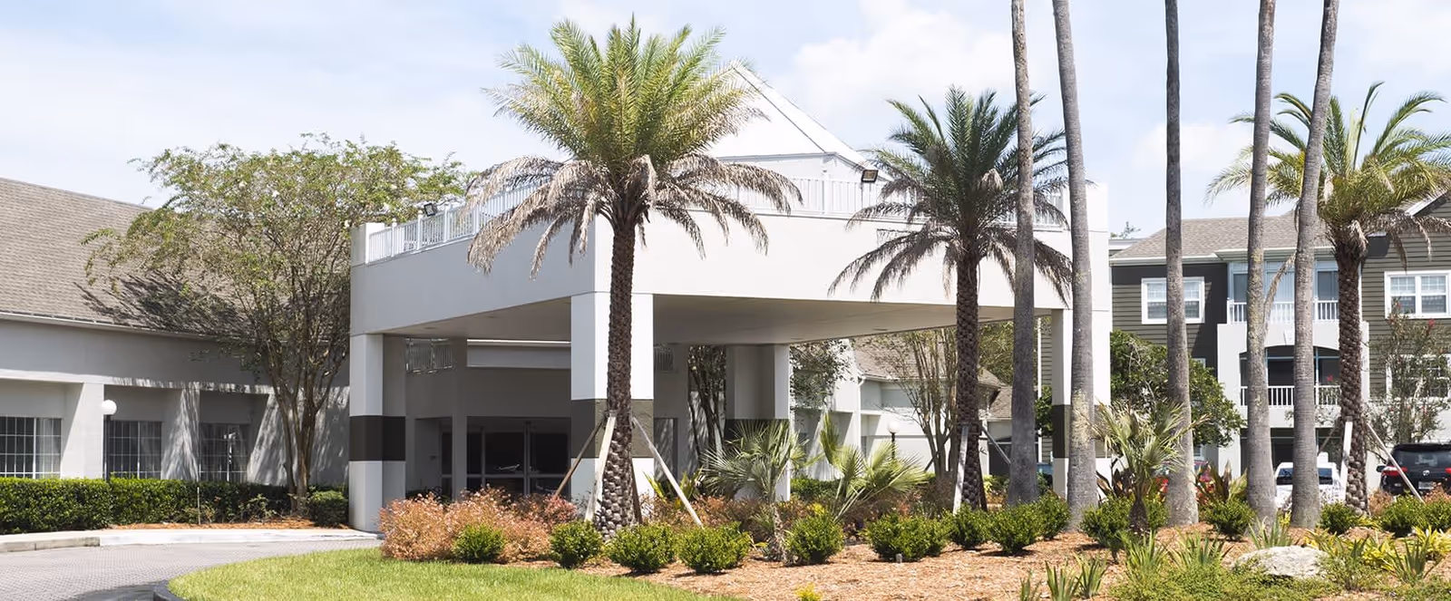 Exterior view of Brookdale Southside senior living facility showing the entrance area with a covered drop-off, surrounded by palm trees and landscaped greenery under a partly cloudy sky.
