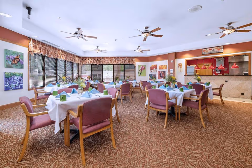 A bright dining room with multiple tables covered in white tablecloths, each set with blue napkins and green mugs. The room has large windows with blinds and valances, ceiling fans, and colorful artwork on the walls. There is a serving area with red pendant lights and a tiled counter.