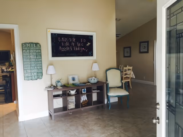 Interior view of an assisted living facility entrance area with beige walls and tiled floor. A small console table with decorative items and two lamps is against the wall, next to a green cushioned chair. Above the table is a framed chalkboard sign that reads 'Welcome to Path of Life Assisted Living.' To the left, a doorway leads to a kitchen area, and a decorative sign with inspirational text is mounted on the wall. A hallway with framed pictures is visible in the background.