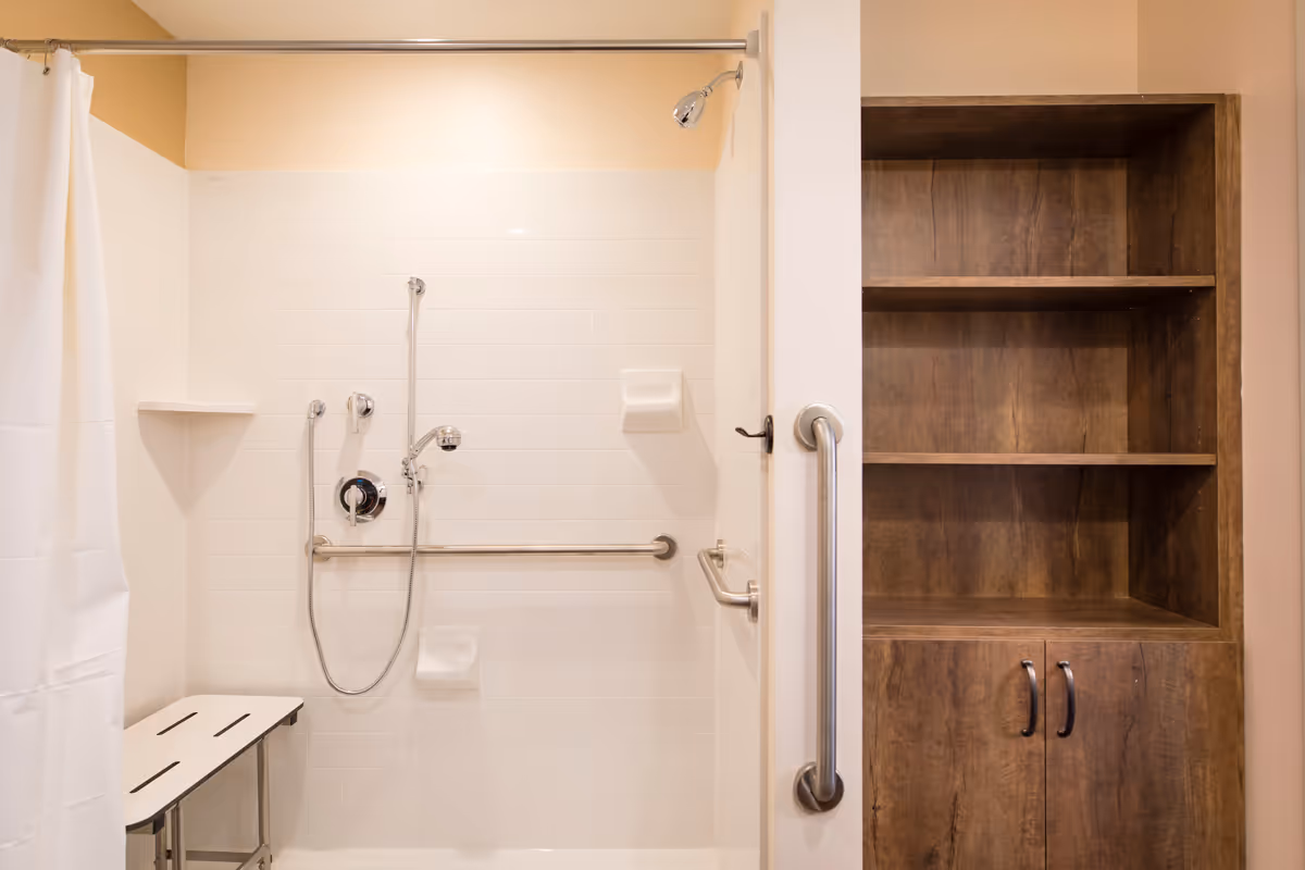 Accessible shower area with white tiled walls, a handheld showerhead, grab bars, a foldable shower bench, and a wooden storage cabinet with shelves and closed compartments next to the shower.