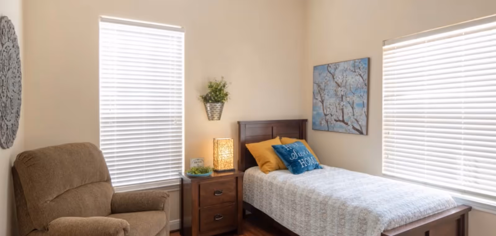 A cozy bedroom with a single wooden bed covered in a white quilt and two pillows, one mustard yellow and one blue with the words 'Home Sweet Home'. Next to the bed is a wooden nightstand with a decorative lamp, a small plant, and a framed sign. There is a brown upholstered armchair to the left, two windows with white blinds, a wall-mounted plant, a decorative wall piece, and a painting of tree branches with white flowers on the wall.