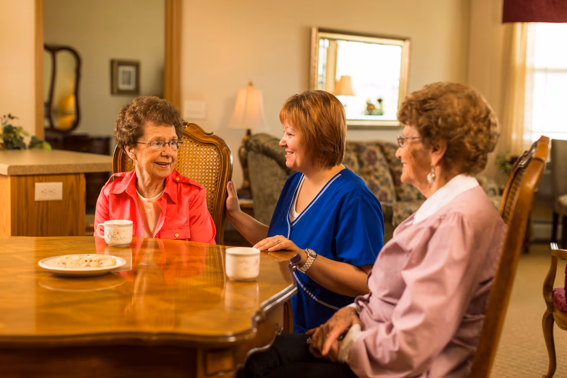 Two elderly women and a caregiver sitting around a wooden table in a cozy living room, smiling and engaging in conversation. There are cups and a plate of cookies on the table, with a lamp and a mirror in the background.