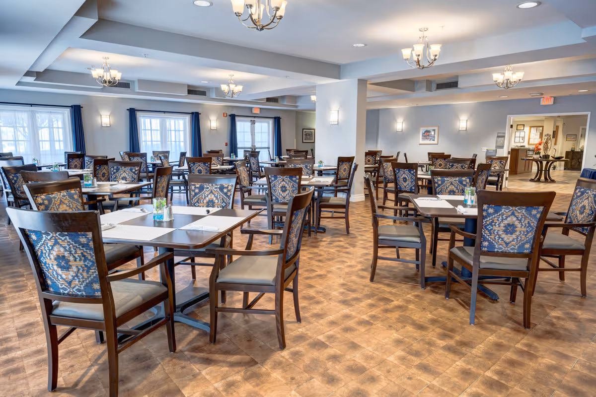 A spacious dining room with multiple wooden tables and chairs featuring patterned upholstery. The room has large windows with blue curtains, light-colored walls, and ceiling lights with chandeliers. The floor has a wood-like finish, and the room appears clean and well-lit.