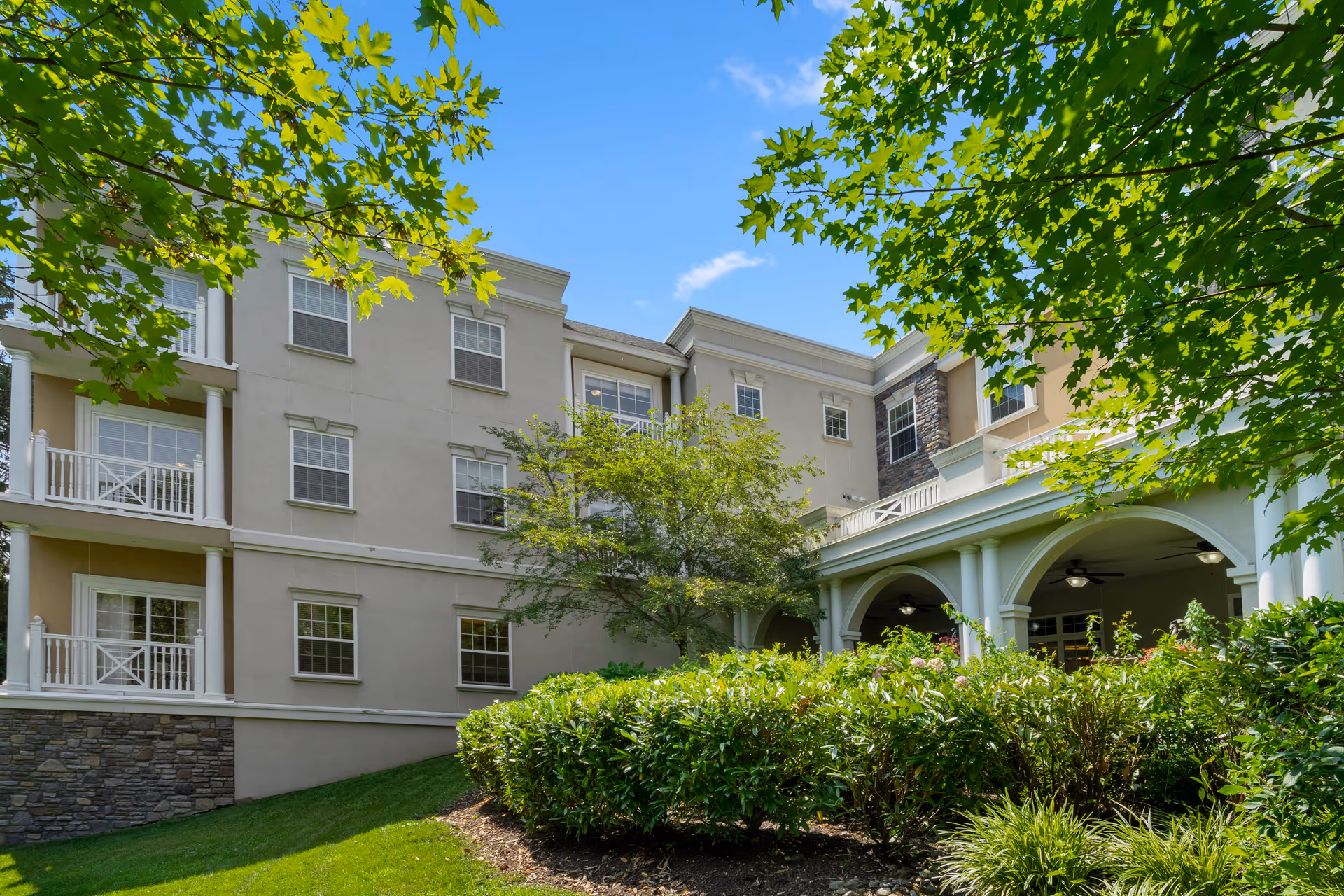 Exterior view of a multi-story senior living facility building with beige walls, white railings, and stone accents. The foreground features green bushes, trees, and a well-maintained lawn under a clear blue sky.