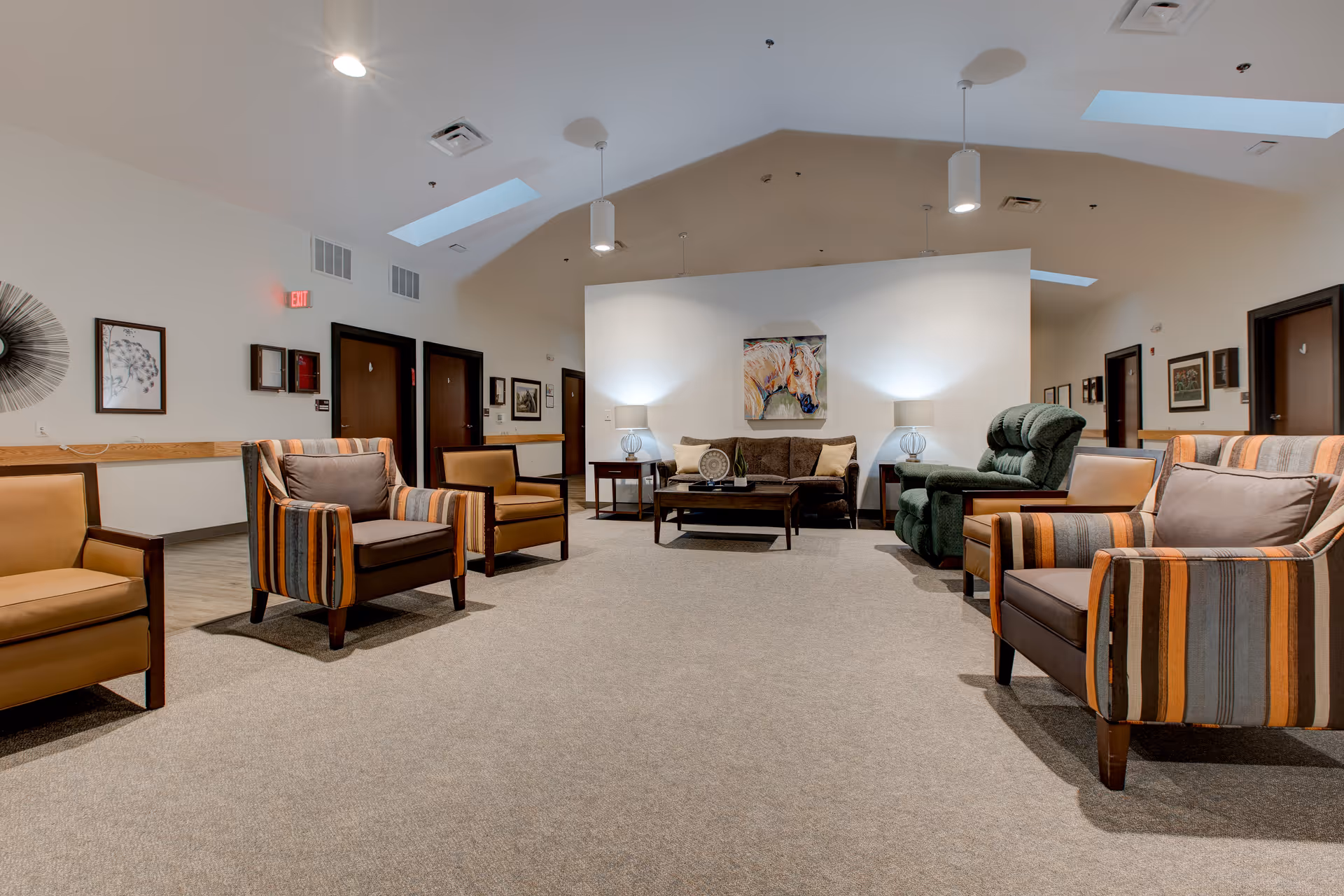 A spacious senior living lounge with multiple upholstered armchairs and sofas arranged around a coffee table beneath skylights and ceiling lights.