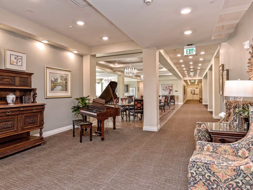 Interior view of a senior living facility hallway with a grand piano and bench on the left, antique wooden cabinet, framed artwork on the walls, and a seating area with patterned armchairs and a side table with a lamp on the right. The hallway leads to a dining area with tables and chairs, illuminated by chandeliers and recessed ceiling lights.
