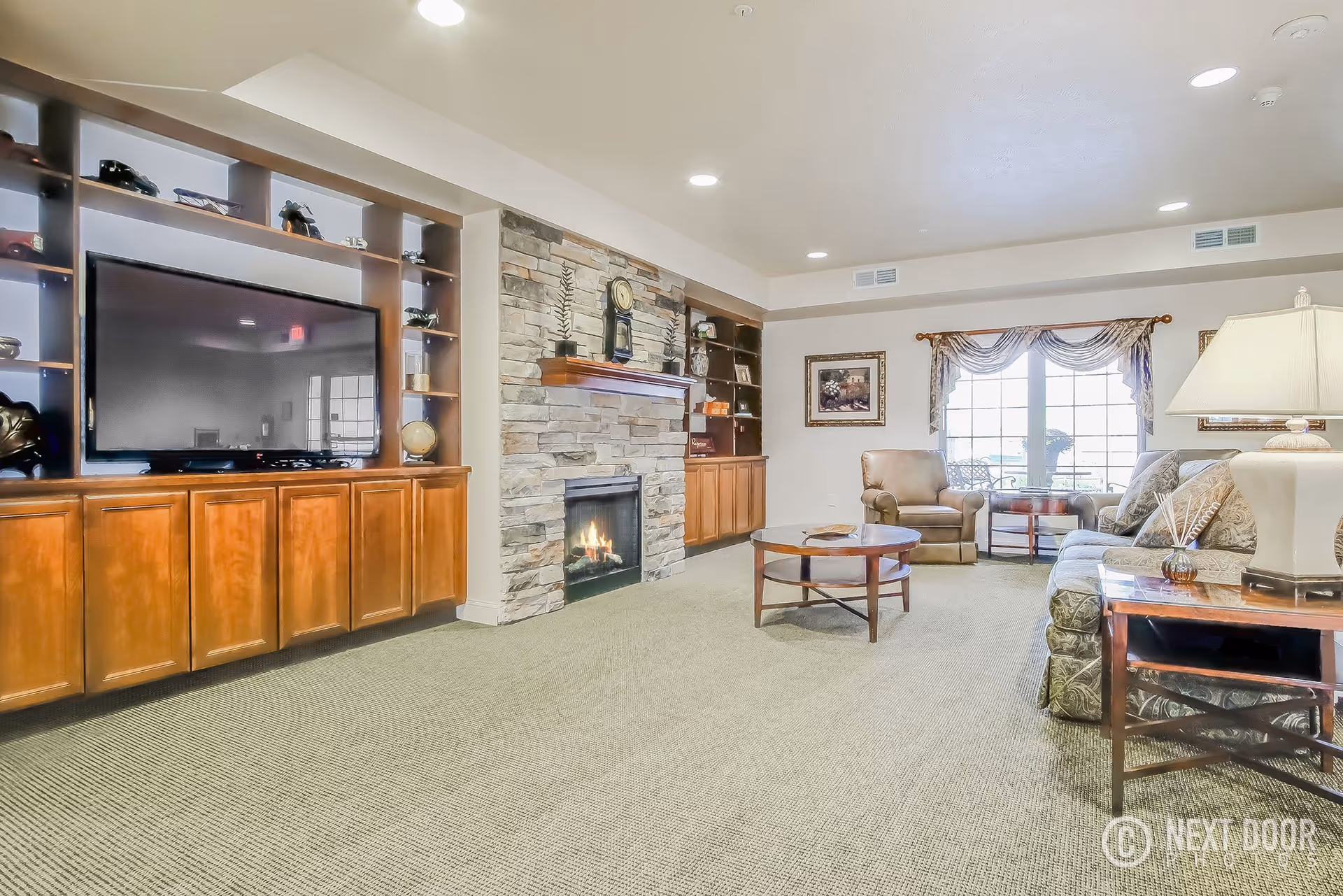 A spacious living room with a large flat-screen TV mounted on a wooden entertainment center, a stone fireplace with a clock on the mantel, a round wooden coffee table, an armchair, a sofa with patterned cushions, a side table with a lamp, and a large window with decorative curtains letting in natural light.