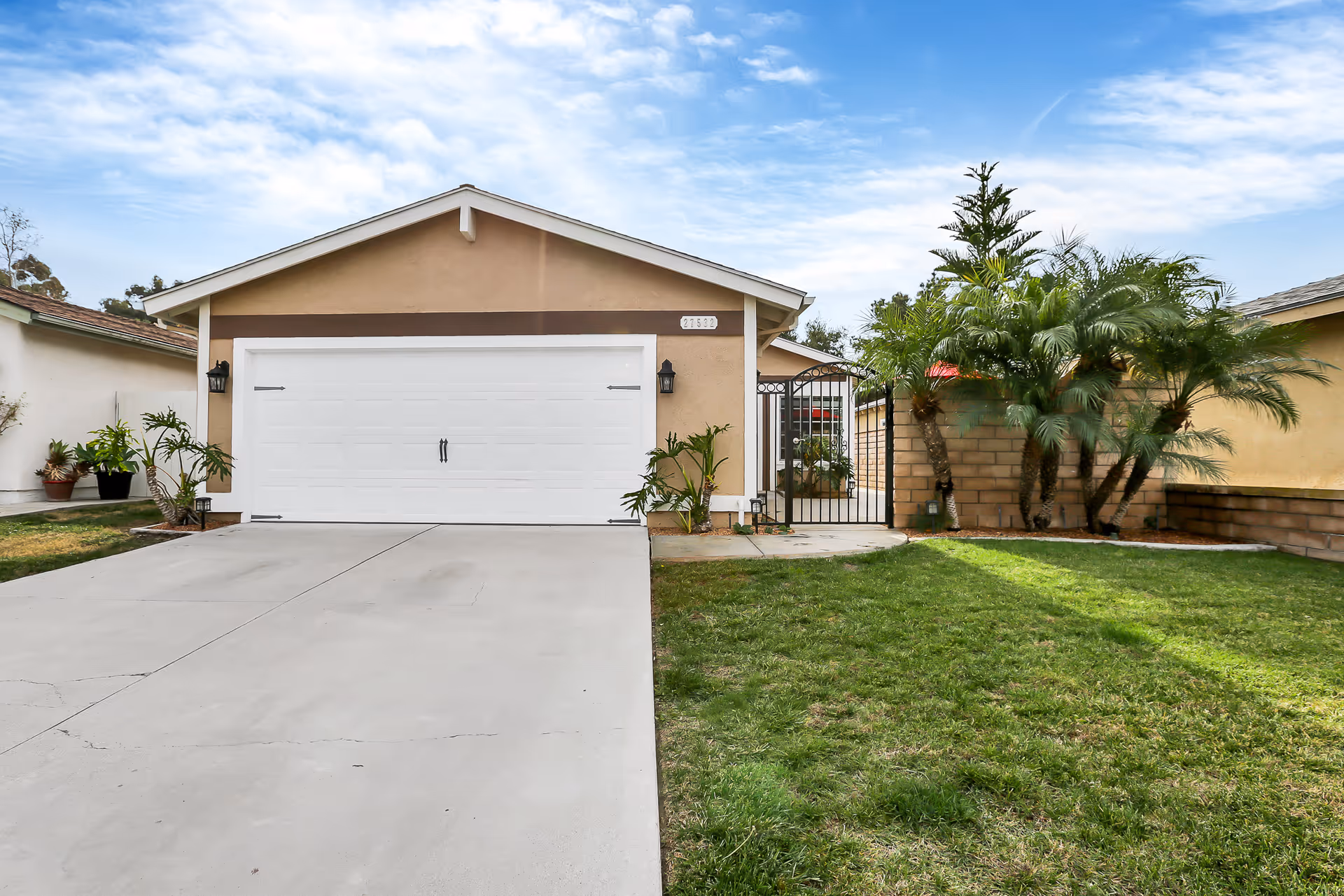 Front view of a single-story house with a white garage door, concrete driveway, green lawn, and a gated side entrance flanked by palm trees.