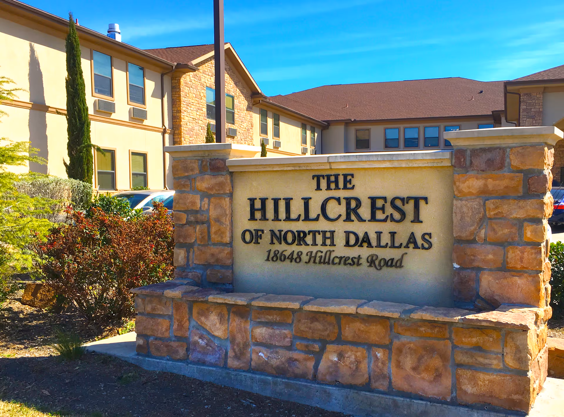 Stone sign at the entrance of The Hillcrest of North Dallas facility with the address 18648 Hillcrest Road, surrounded by bushes and a two-story building in the background under a clear blue sky.