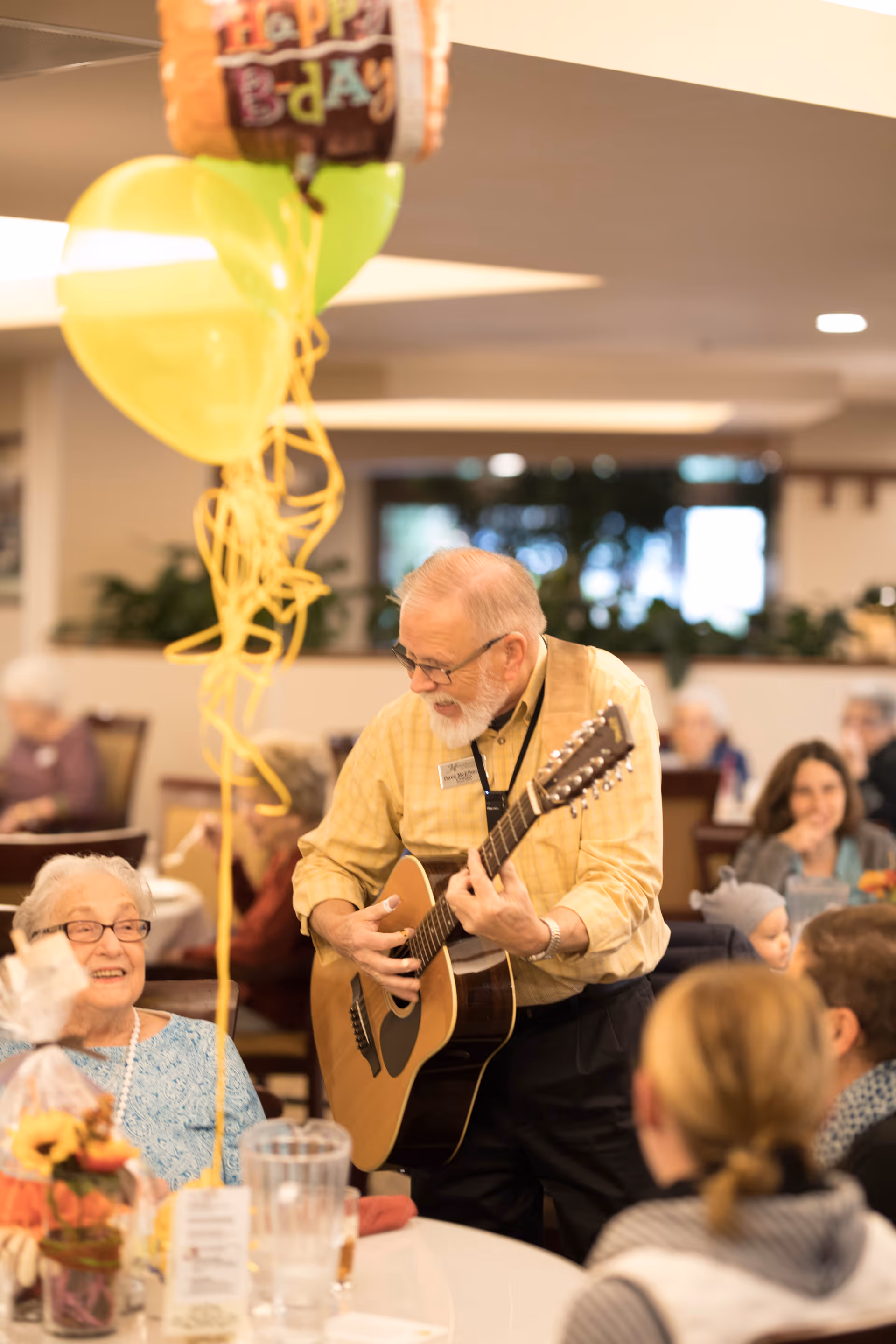 An elderly man playing an acoustic guitar and singing to a group of seated seniors in a communal dining area decorated with balloons, including one that says 'Happy B-day'.