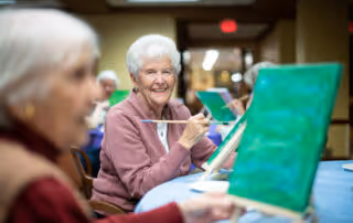 Elderly women sitting at a table engaged in painting on canvases, smiling and enjoying an art activity in a well-lit indoor setting.