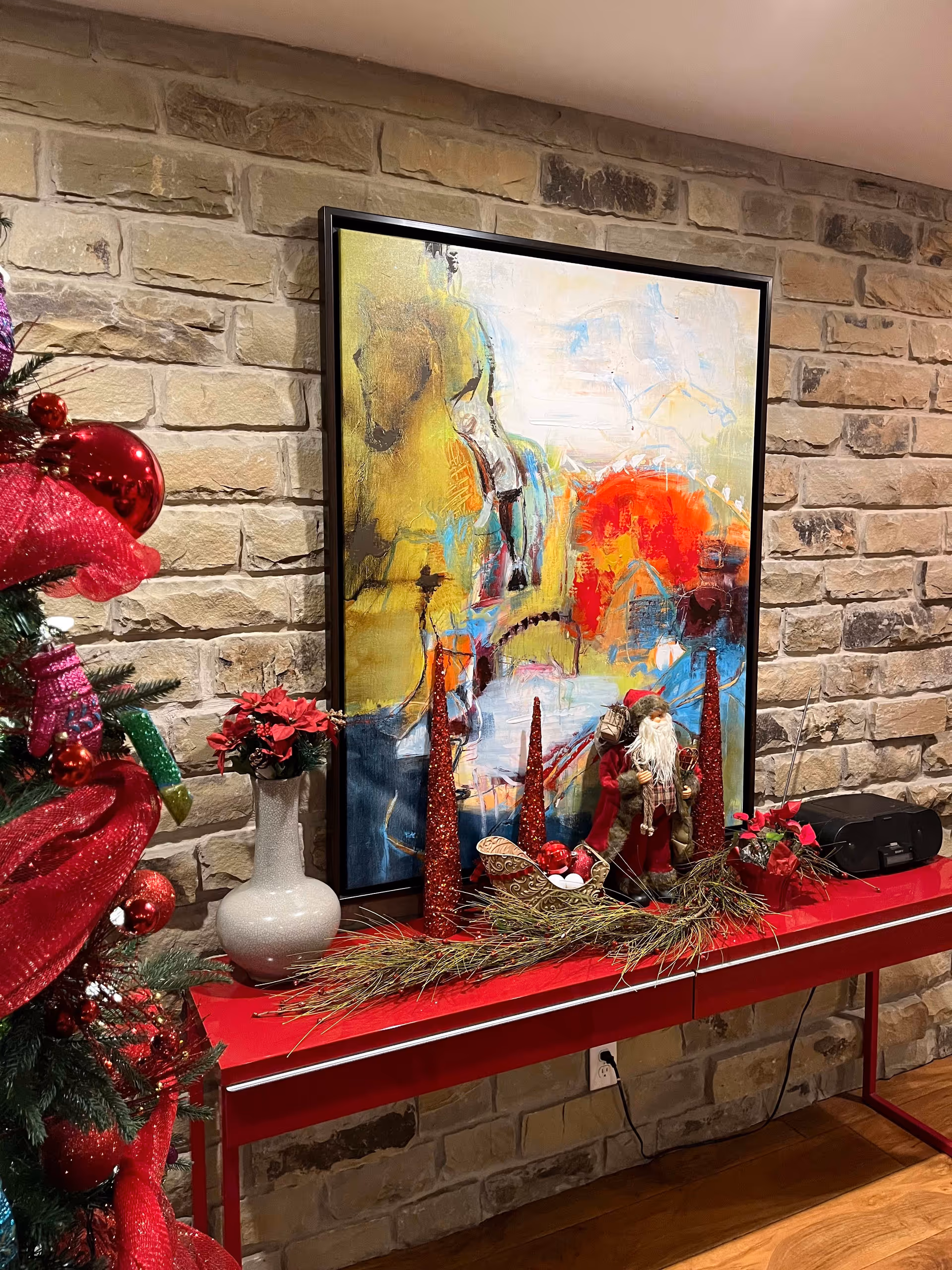 A red console table against a stone wall decorated with Christmas-themed items including a Santa figurine, red glittery cone-shaped ornaments, a small sleigh with ornaments, a white vase with red flowers, and greenery. To the left, part of a decorated Christmas tree with red ribbons and ornaments is visible. Above the table hangs a large abstract painting with bright colors including yellow, red, blue, and white.