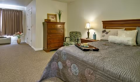 Well-lit senior living bedroom with a bed, nightstand and lamp, dresser, upholstered chair, and a window with curtains.
