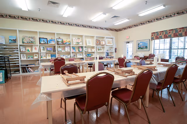 A bright and spacious activity room with tables covered in plastic tablecloths and wooden easels set up for painting or drawing. Red cushioned chairs surround the tables. The back wall features shelves filled with various paintings and art supplies. Large windows with patterned valances allow natural light to enter the room.