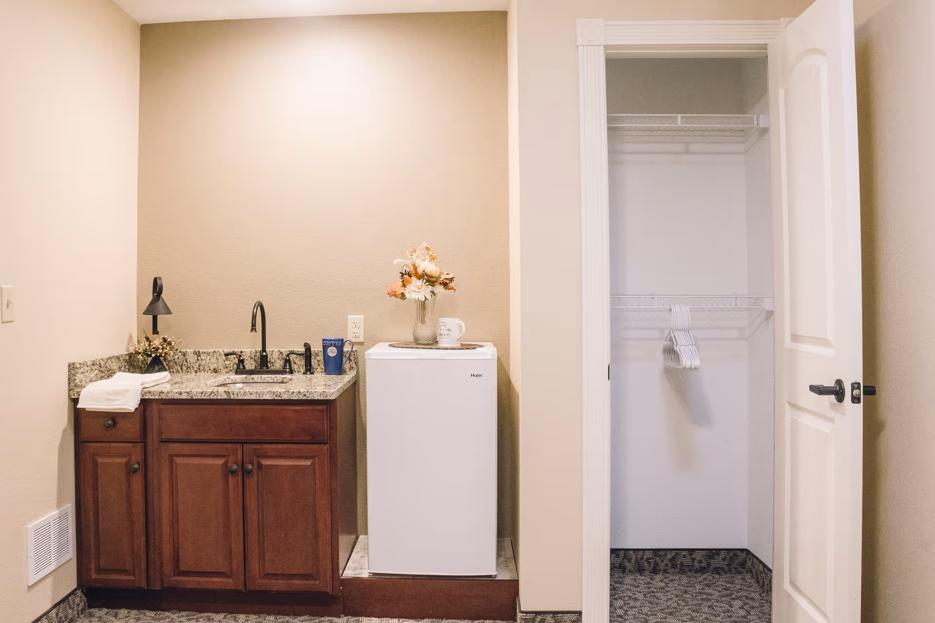 Small kitchenette with a granite sink and mini fridge beside an open closet with hangers.