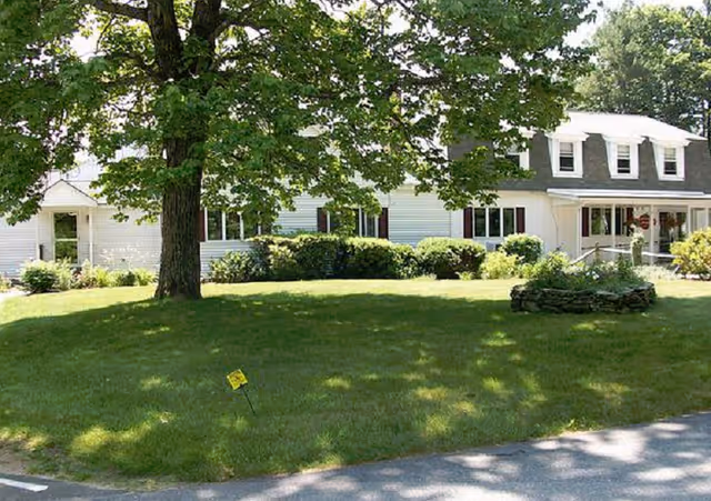 Front exterior of a white two-story building with dormer windows, a large shaded lawn, shrubs, and a tree.