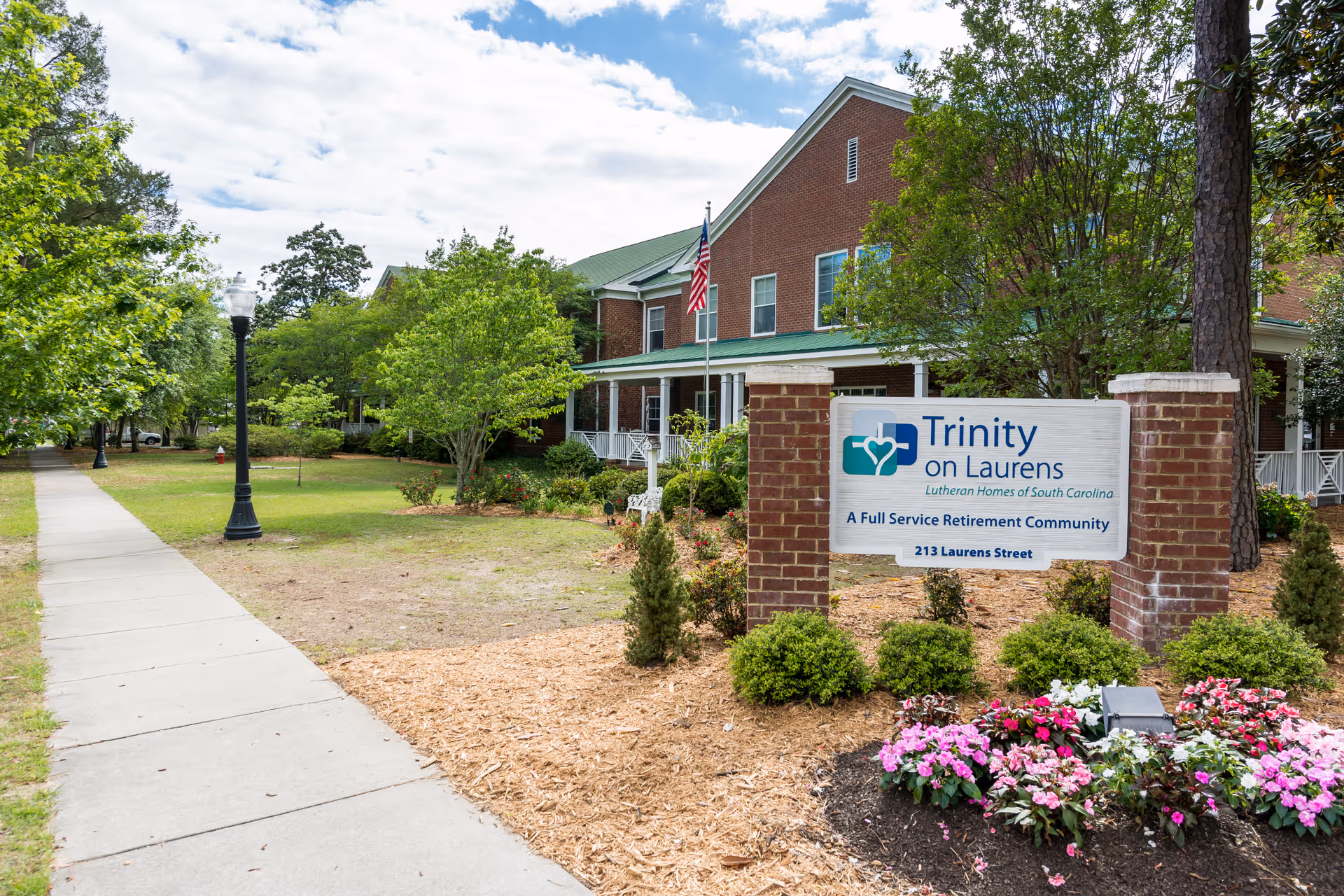 Exterior view of Trinity on Laurens, a full-service retirement community, showing a brick building with a green roof, an American flag, a sidewalk, trees, and a landscaped garden with flowers and shrubs around the community sign.