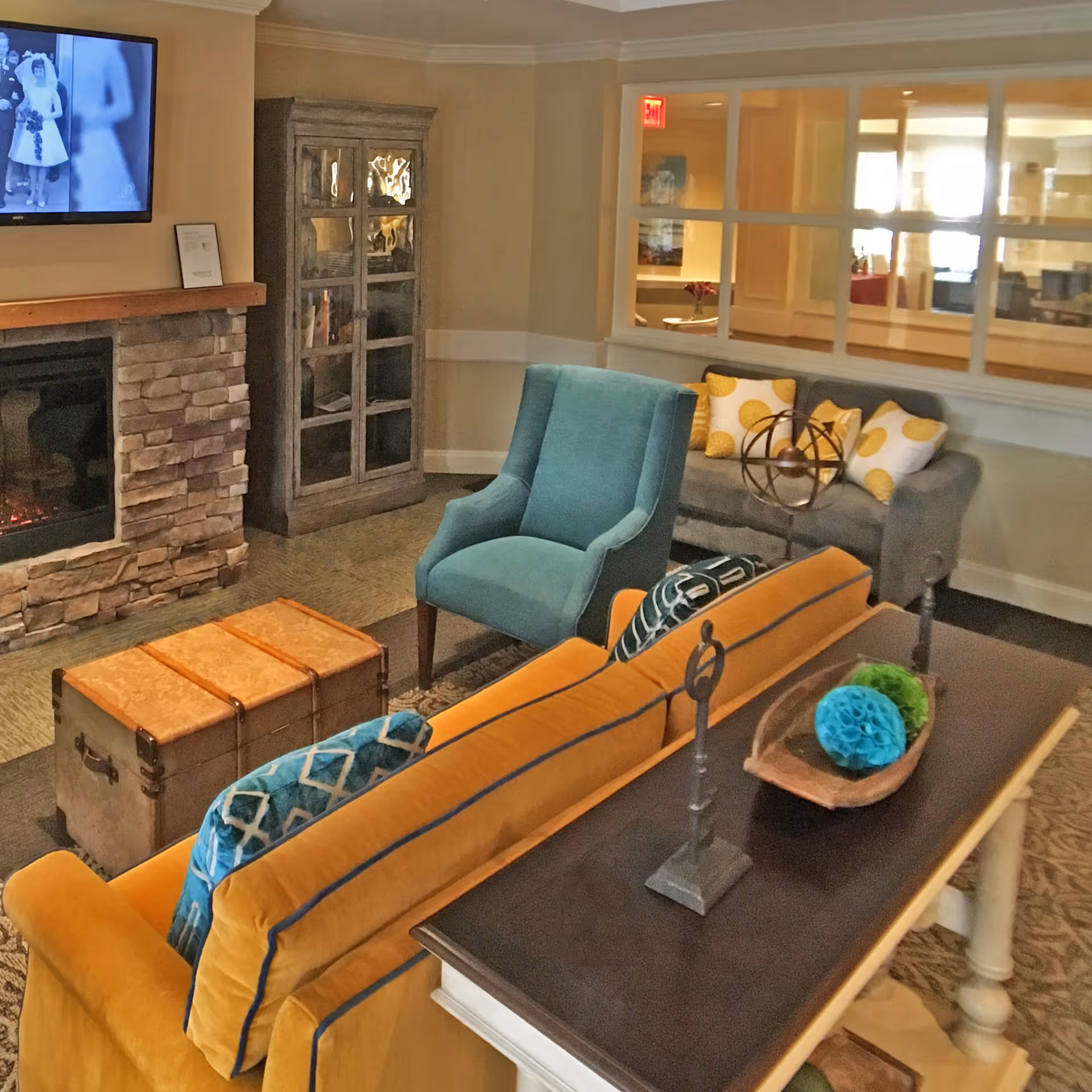 A cozy living room area in a senior living facility featuring a mustard yellow sofa with blue patterned pillows, a teal armchair, and a gray loveseat with yellow and white polka dot pillows. There is a stone fireplace with a mounted TV above it displaying a black and white image. A wooden cabinet with glass doors stands next to the fireplace. A wooden trunk serves as a coffee table, and a console table behind the sofa holds decorative items including a wooden bowl with blue and green paper decorations and two metal candle holders. The room has beige walls and carpeted flooring, with a windowed partition showing an adjacent room.