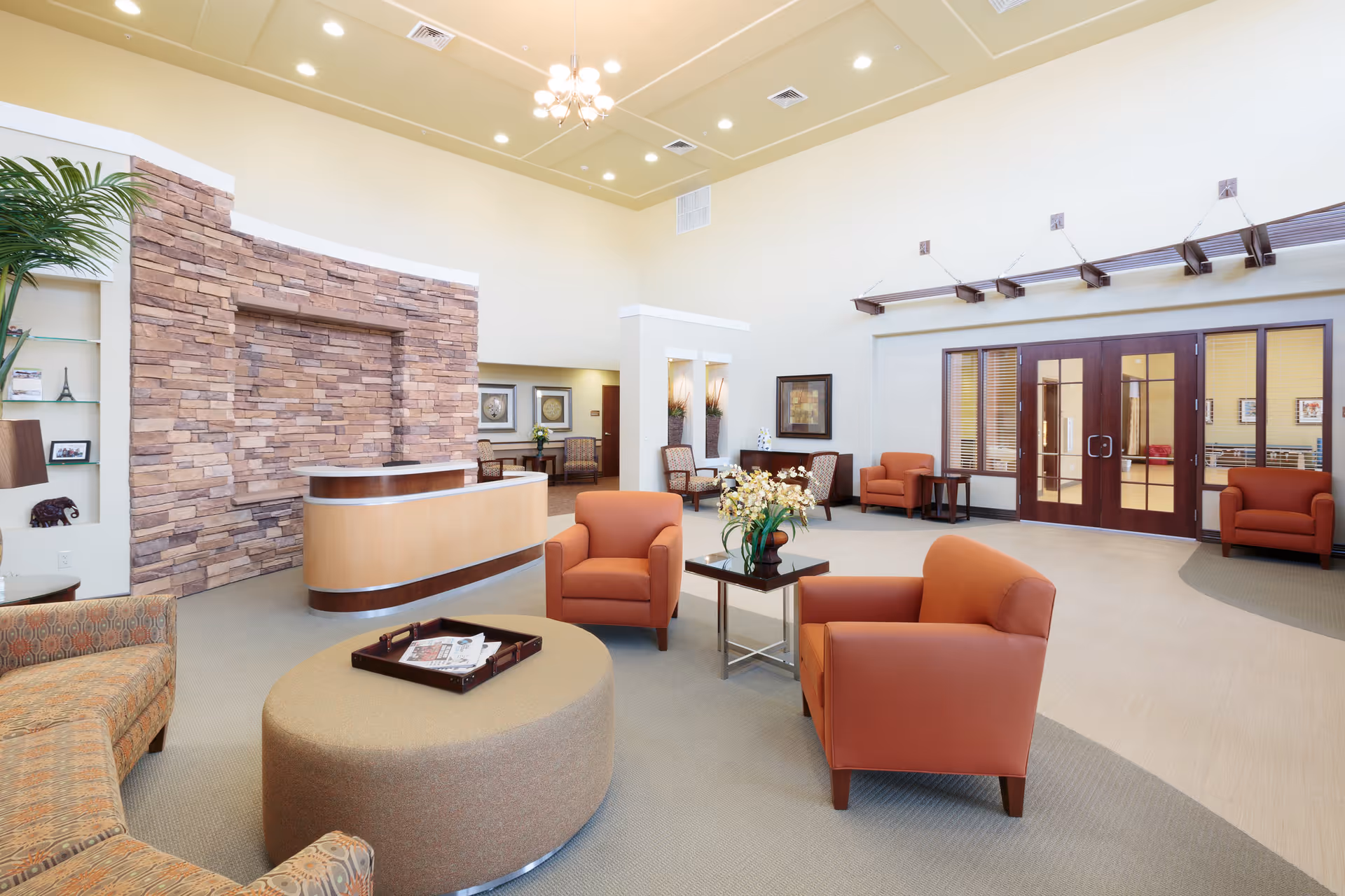 A spacious and well-lit senior living facility common area with comfortable orange armchairs, a round ottoman with a tray of magazines, a stone accent wall behind a curved reception desk, and large wooden double doors with glass panels. The room features high ceilings with modern light fixtures and decorative plants.