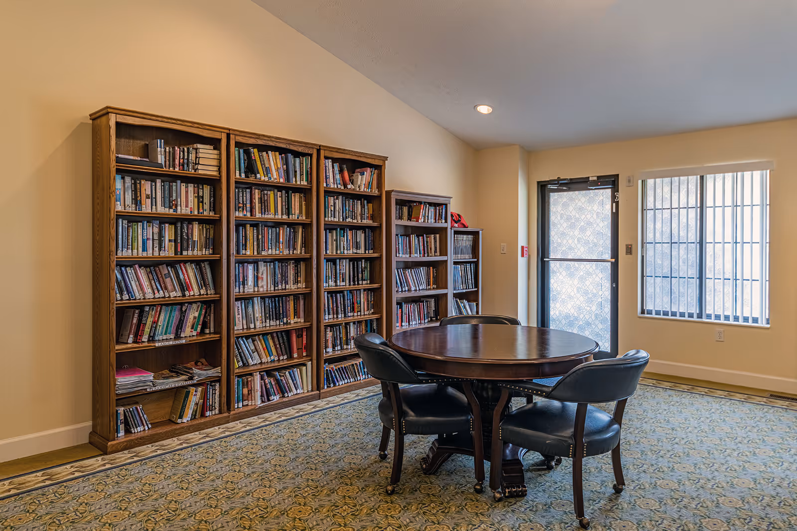 A cozy reading room with wooden bookshelves filled with books along one wall, a round wooden table with four black cushioned chairs on casters, a patterned carpet, and a door and window with vertical blinds letting in natural light.
