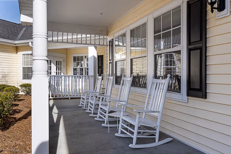 A covered porch area with six white wooden rocking chairs lined up against the yellow siding of a building with multiple windows and black shutters. There are bushes and mulch along the edge of the porch, and the sky is clear and blue.