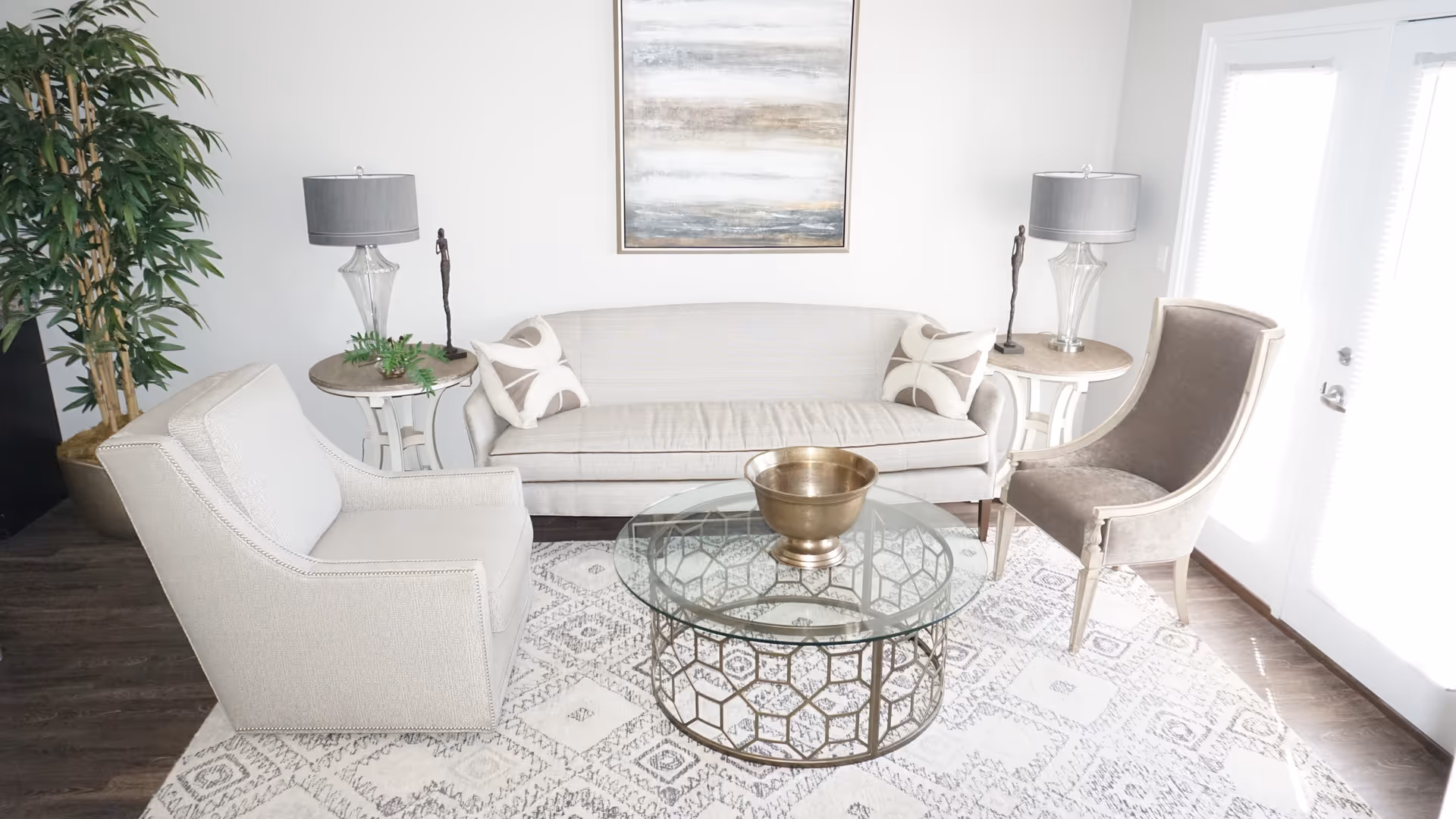 A bright living room with a light gray sofa adorned with two patterned pillows, a beige armchair, and a taupe accent chair. A glass coffee table with a geometric metal base holds a decorative brass bowl. Two side tables with glass lamps and small sculptures flank the sofa. A large abstract painting hangs on the white wall behind the sofa, and a tall potted plant is visible on the left. The room has wooden flooring and a patterned area rug, with double glass doors on the right letting in natural light.