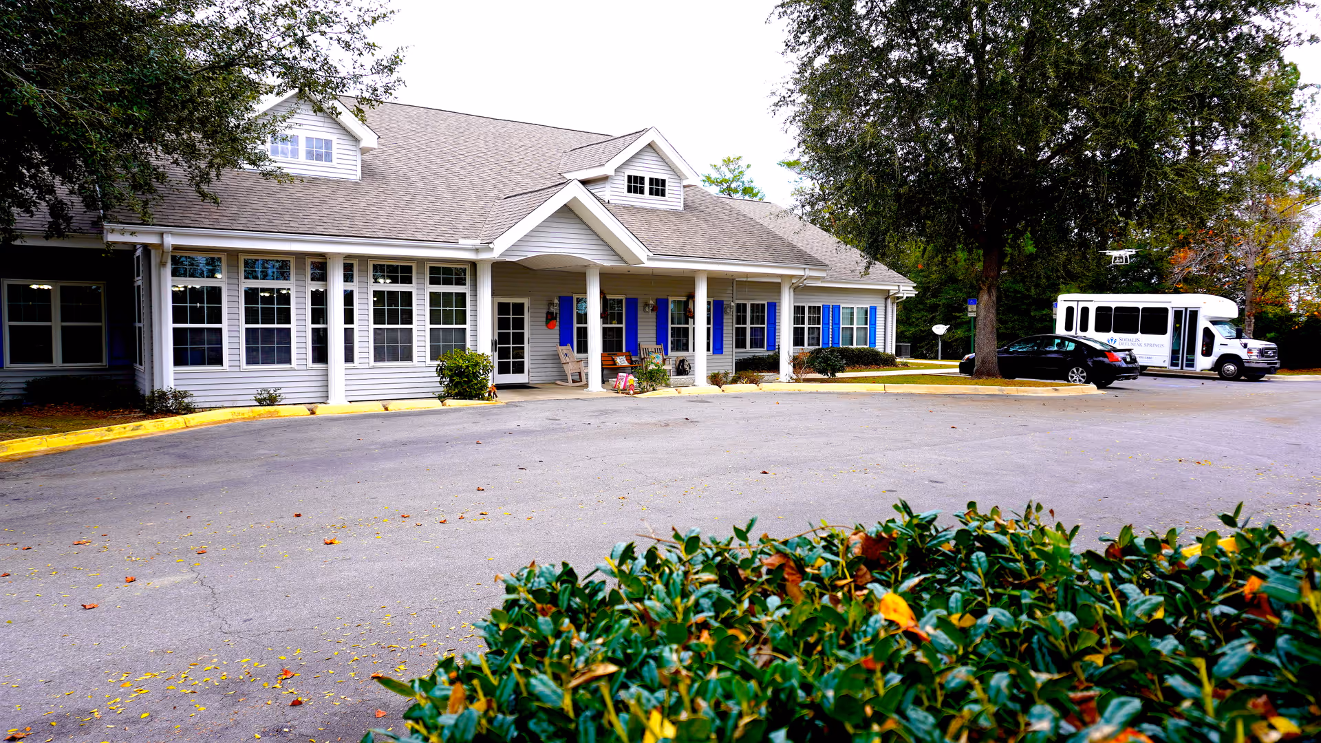 Exterior view of a single-story senior living facility building with a covered entrance, several windows, and blue shutters. There is a paved driveway in front with some parked vehicles, including a white shuttle bus. Green shrubs and trees surround the area.