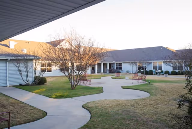 Outdoor courtyard area of a senior living facility with paved walkways, leafless trees, benches, and a single-story building surrounding the courtyard under a clear sky.