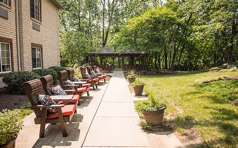 Outdoor patio area at Greenfield Senior Living at Graysonview with wooden chairs featuring red cushions and patterned pillows lined up along a concrete walkway. There are potted plants along the path and a wooden gazebo surrounded by trees and greenery in the background.