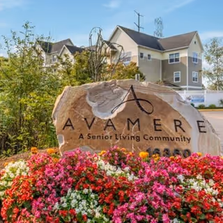 Large stone sign with the text 'AVAMERE A Senior Living Community' surrounded by colorful flowers and greenery, with a multi-story residential building in the background under a clear blue sky.