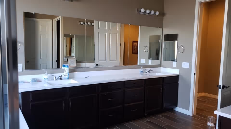 A spacious bathroom with a long white countertop featuring two sinks and dark wooden cabinets underneath. Above the countertop is a large mirror with a row of lights mounted above it. The bathroom has beige walls and a tiled floor. There are two open doors visible, one leading to another room and the other to a hallway.