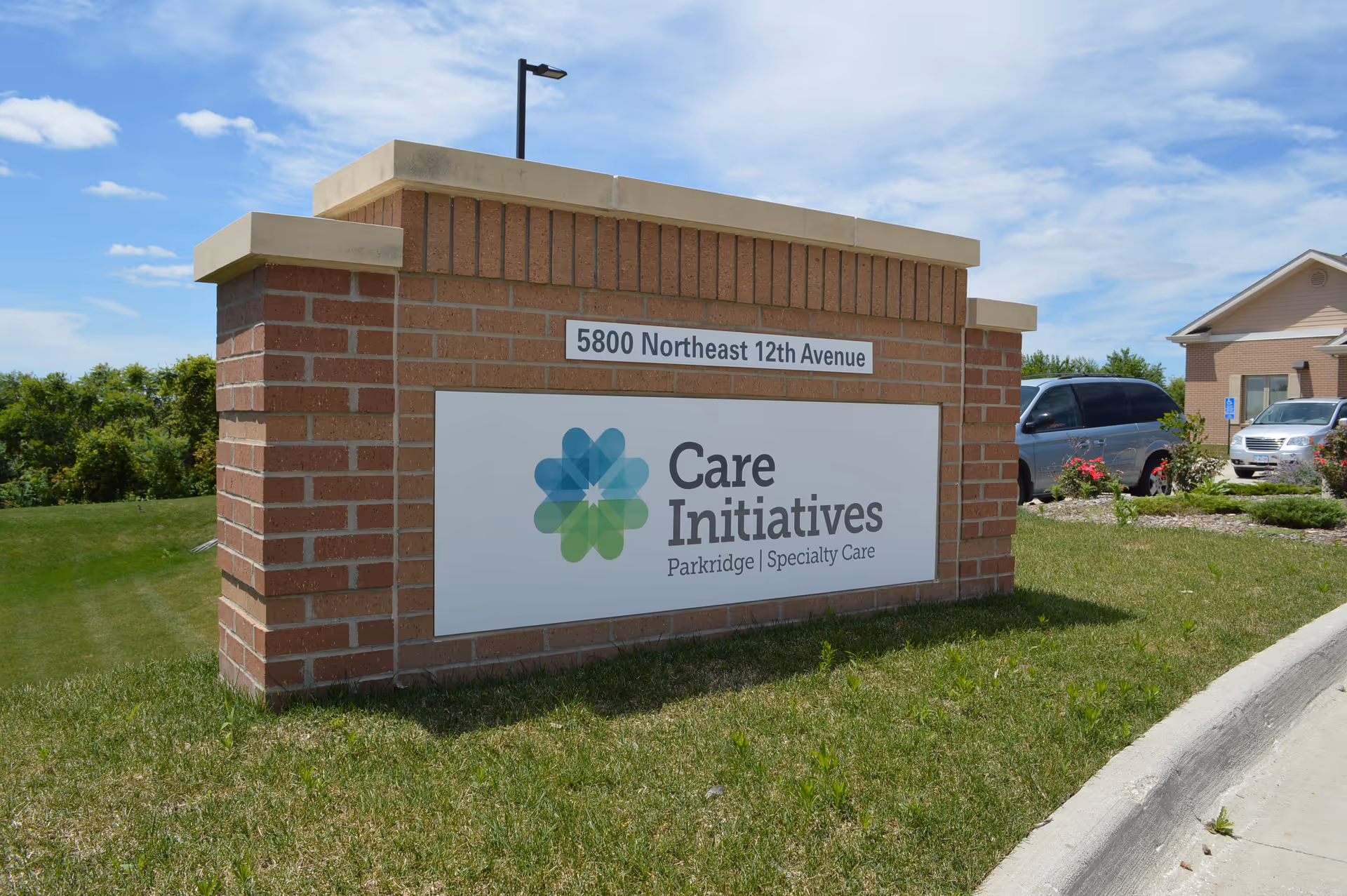 A brick sign outside on a grassy area with the address 5800 Northeast 12th Avenue displayed above a logo and text that reads Care Initiatives Parkridge | Specialty Care. There are cars and a building visible in the background under a partly cloudy sky.