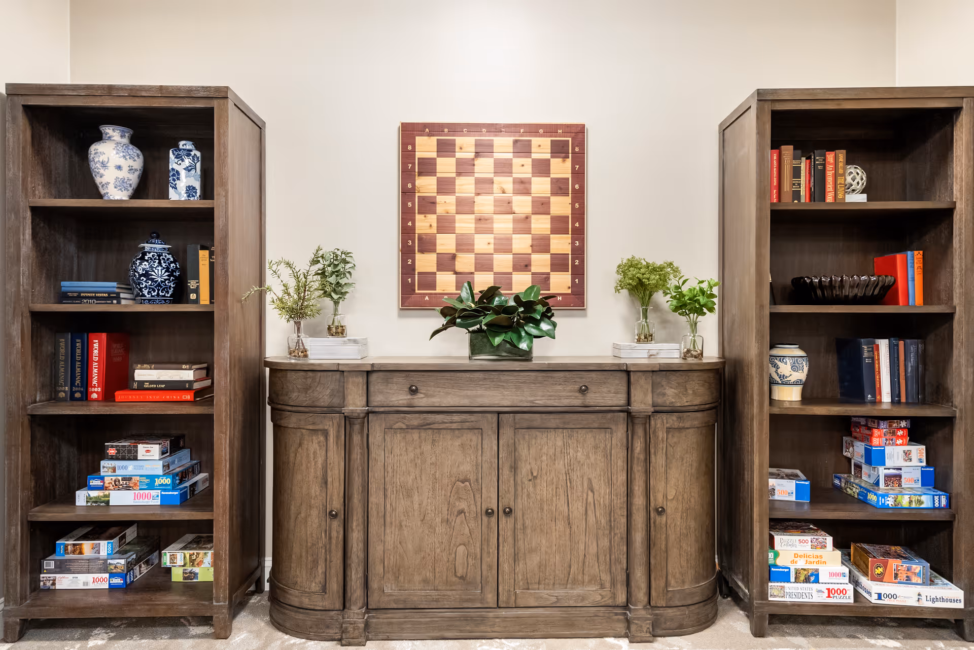 Wooden cabinet flanked by two bookcases holding books, puzzles and decorative vases, with plants on the cabinet and a wall-mounted chessboard above.
