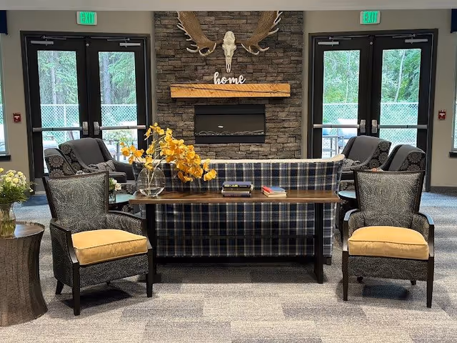 A cozy senior living common area with a stone fireplace featuring a mounted moose skull and antlers above a wooden mantel with the word 'home'. In front of the fireplace is a plaid sofa with a wooden console table behind it, decorated with a vase of yellow flowers and some books. Two patterned armchairs with yellow cushions are positioned facing the sofa. Large windows and double doors with exit signs provide natural light and a view of greenery outside.