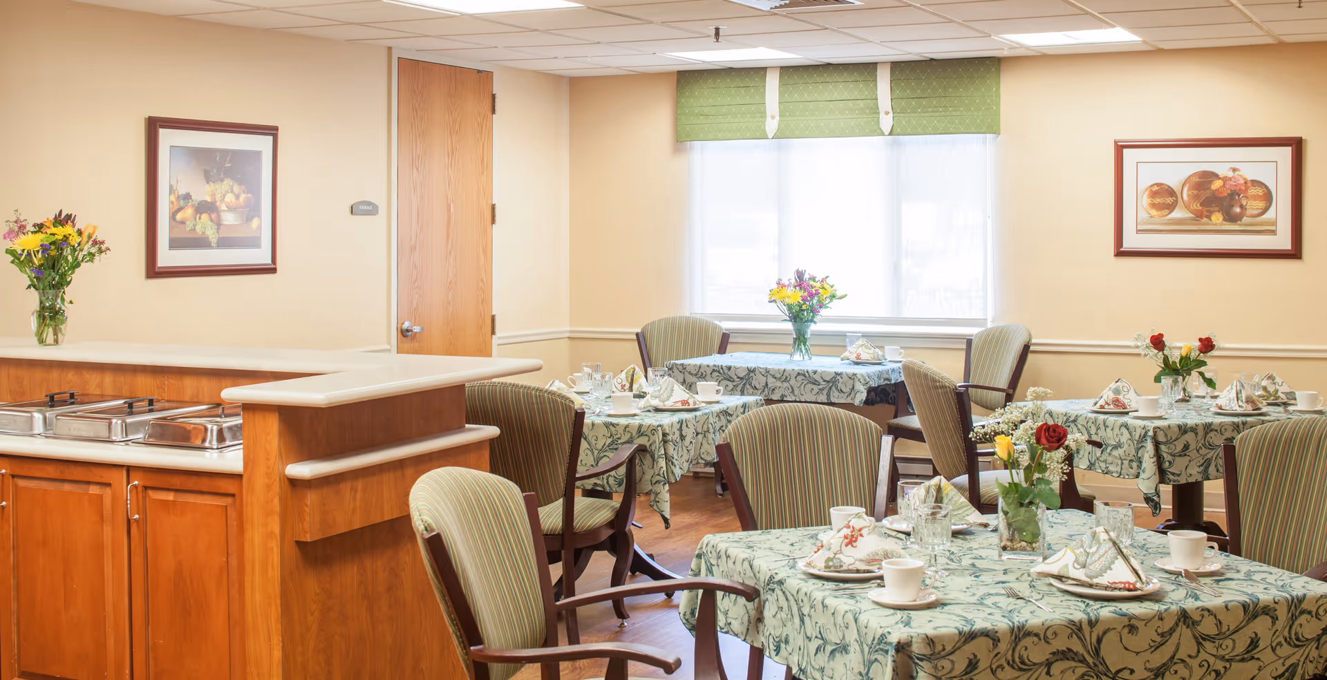 Bright dining room with several tables set with tablecloths, place settings, floral centerpieces, upholstered chairs, and a serving counter.
