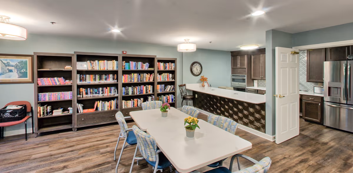 Community common room with a long table and chairs, bookshelves along the wall and a kitchenette/bar area in the background.
