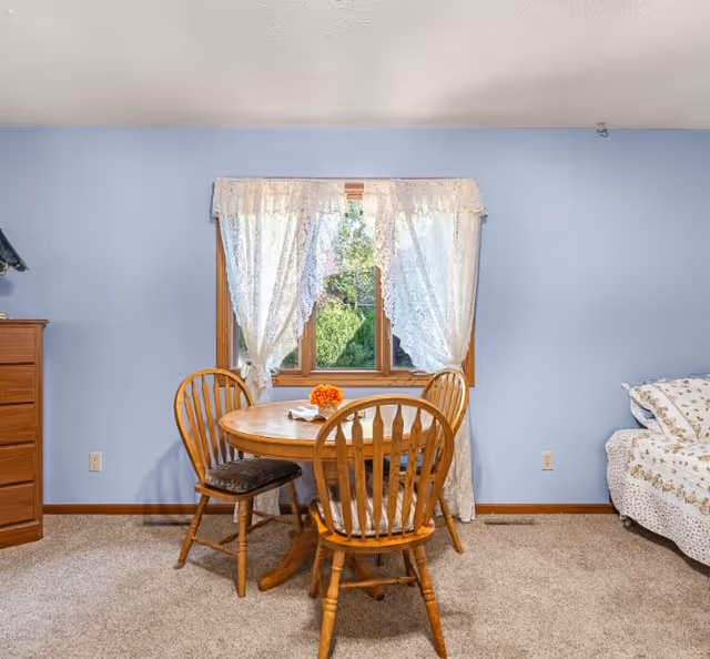 Wooden round dining table with three chairs set in front of a window with lace curtains in a blue-walled room.