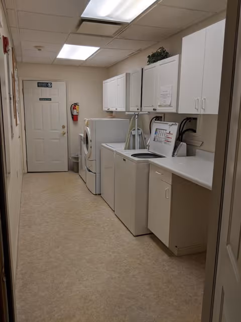 A laundry room with a washing machine and dryer side by side along the right wall. Above the machines are white cabinets for storage. There is a countertop next to the washing machine with an ironing board leaning against the wall. The room has beige flooring and white walls with fluorescent ceiling lights. A door is visible at the end of the room with a fire extinguisher mounted on the wall nearby.