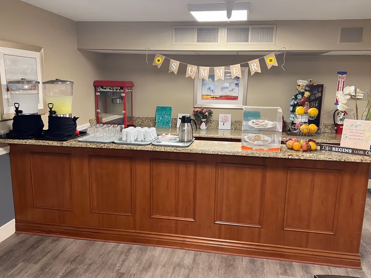 A refreshment counter in a senior living facility with two beverage dispensers containing water and lemonade, a popcorn machine, stacks of glasses and cups, a coffee pot, a display case with cookies, a fruit stand with lemons and apples, and decorative items including a 'SUMMER' banner, paintings, flowers, and a small chef figurine.