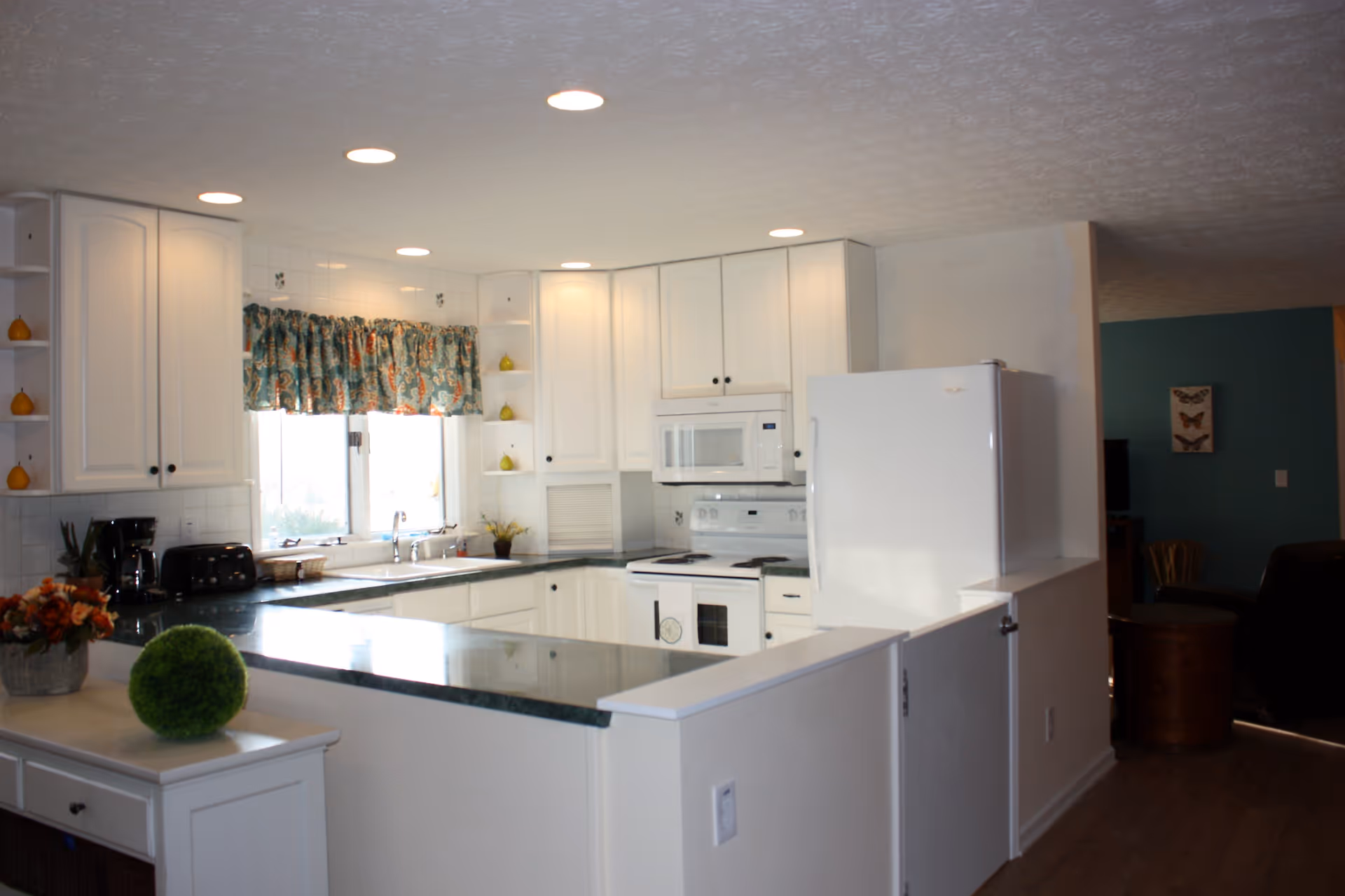 Bright kitchen with white cabinets, a white refrigerator, stove, microwave, and a green countertop. There is a window with patterned curtains above the sink, and various small decorative items on shelves and counters. The kitchen opens into a living area with teal walls and butterfly artwork.