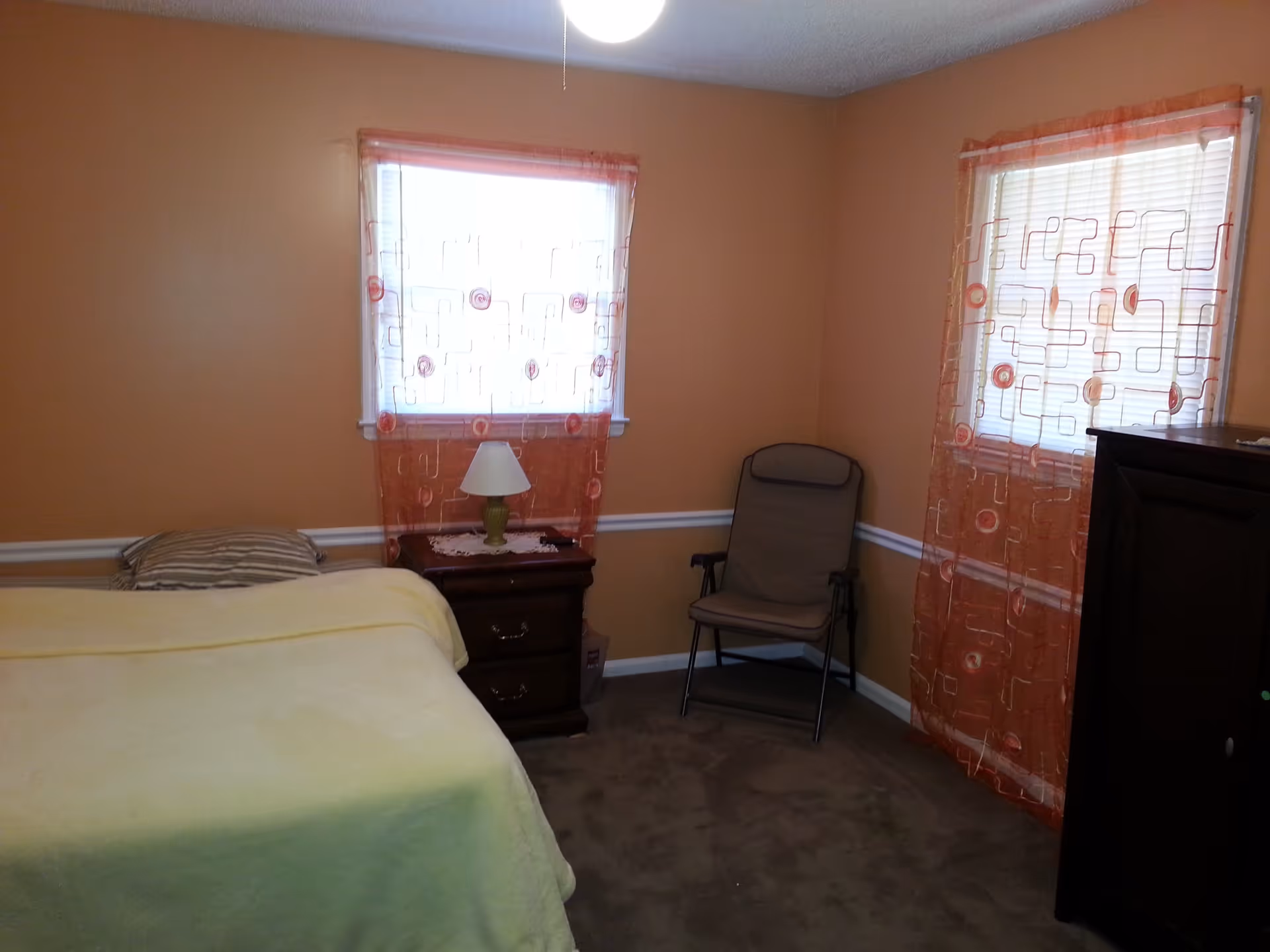 A small bedroom with peach-colored walls and two windows covered with sheer orange curtains featuring a geometric pattern. The room contains a bed with a yellow blanket and striped pillow, a wooden nightstand with a green lamp, a folding chair, and a dark wooden dresser.