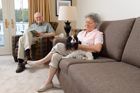An elderly woman sitting on a brown couch petting a black and white dog, while an elderly man sits in a patterned armchair nearby holding a magazine in a well-lit living room with beige carpet and curtains.