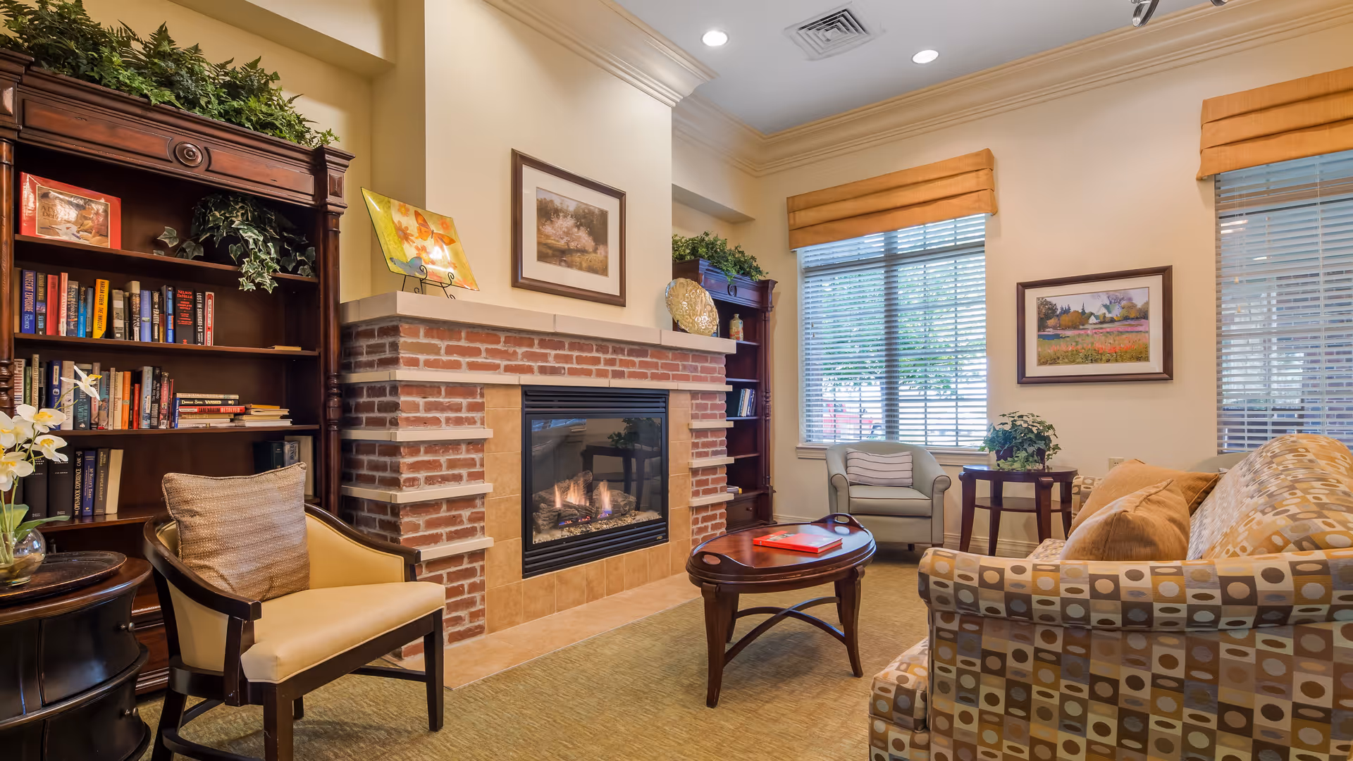A cozy living room area in an assisted living facility featuring a brick fireplace with a fire burning, a wooden bookshelf filled with books and decorative plants, a patterned sofa with cushions, two armchairs, a wooden coffee table with a red book on it, and large windows with blinds and orange valances letting in natural light.