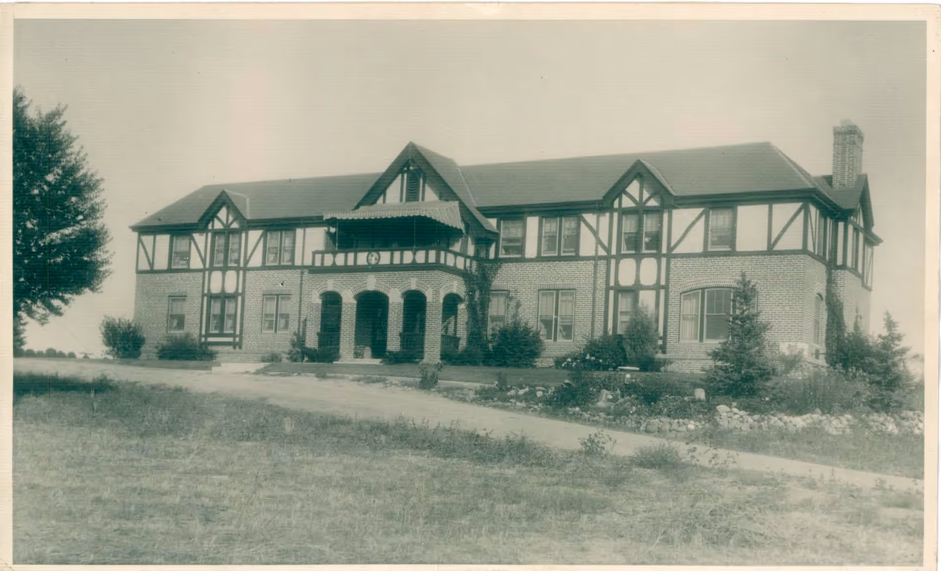 Black and white photograph of a large two-story brick building with Tudor-style architectural elements, including decorative wooden beams and multiple windows. The building has a covered porch with arches and a balcony above it. There is a paved driveway leading up to the entrance and some landscaping with bushes and small trees around the building.