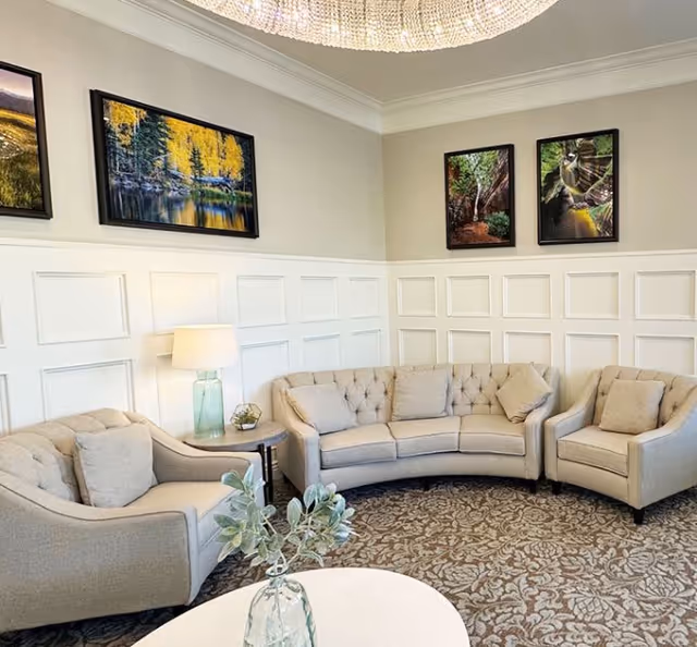A cozy living room area with a curved beige sofa, two matching armchairs, a round white coffee table with a glass vase holding green foliage, a side table with a lamp, and four framed nature photographs on the walls. The room has white paneled walls and a decorative ceiling light fixture.