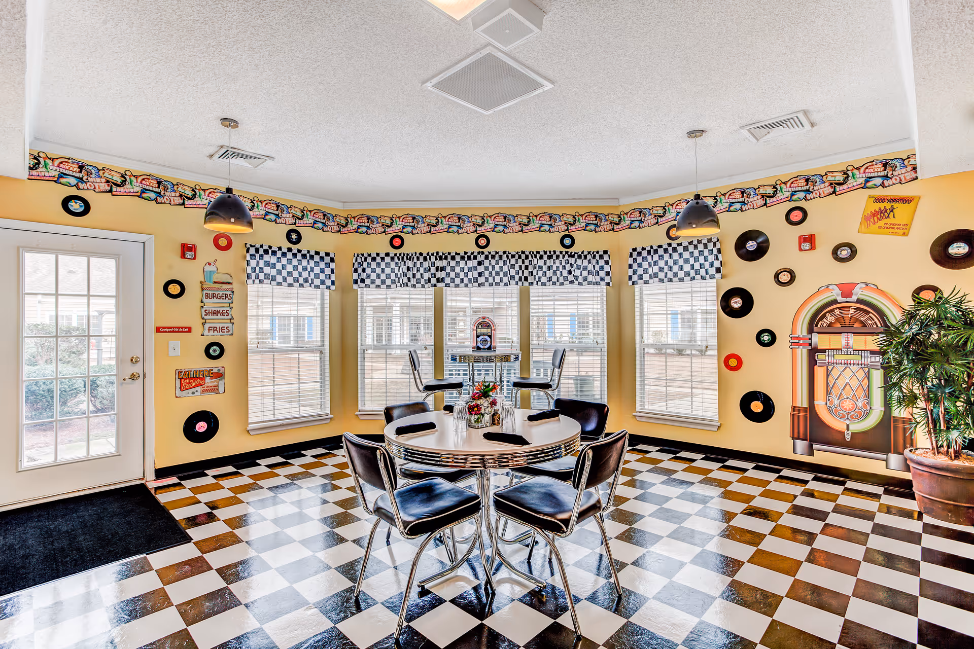A retro-themed dining area with black and white checkered floor tiles and yellow walls decorated with vinyl records and a jukebox mural. The room has a round table with four black chairs in the center and a small table with two chairs by the windows, which have checkered valances. There are two hanging pendant lights and a door leading outside.