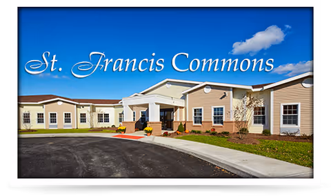 Exterior view of St. Francis Commons Assisted Living Residence building under a blue sky with a few clouds. The building has a beige and light brown facade with multiple windows and a main entrance with a covered porch. There is a paved driveway and some landscaping with grass and small plants.