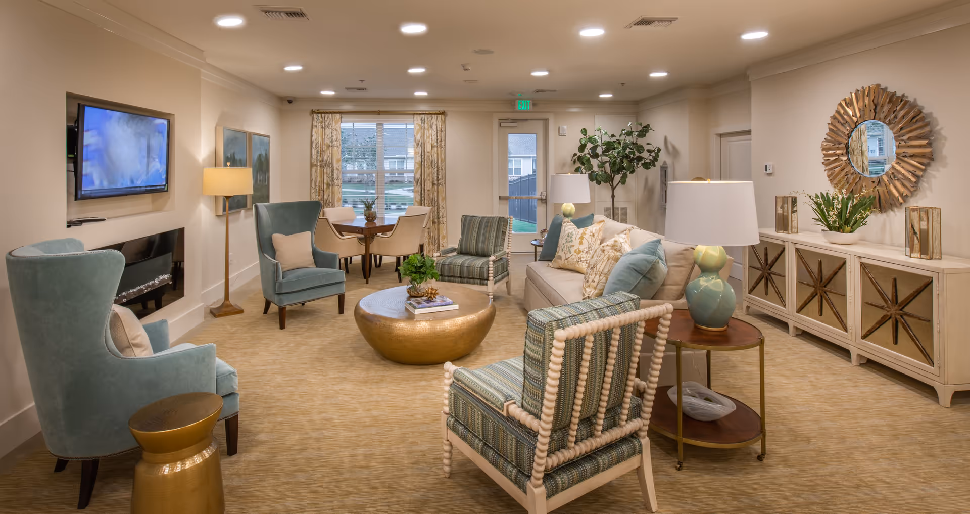 A cozy and well-lit living room area in a senior living facility featuring a beige sofa with decorative pillows, two patterned armchairs, two teal wingback chairs, a round gold coffee table, a side table with a lamp, a wall-mounted TV, a floor lamp, a large window with patterned curtains, a door leading outside, a decorative mirror above a sideboard, and various plants and decor items.