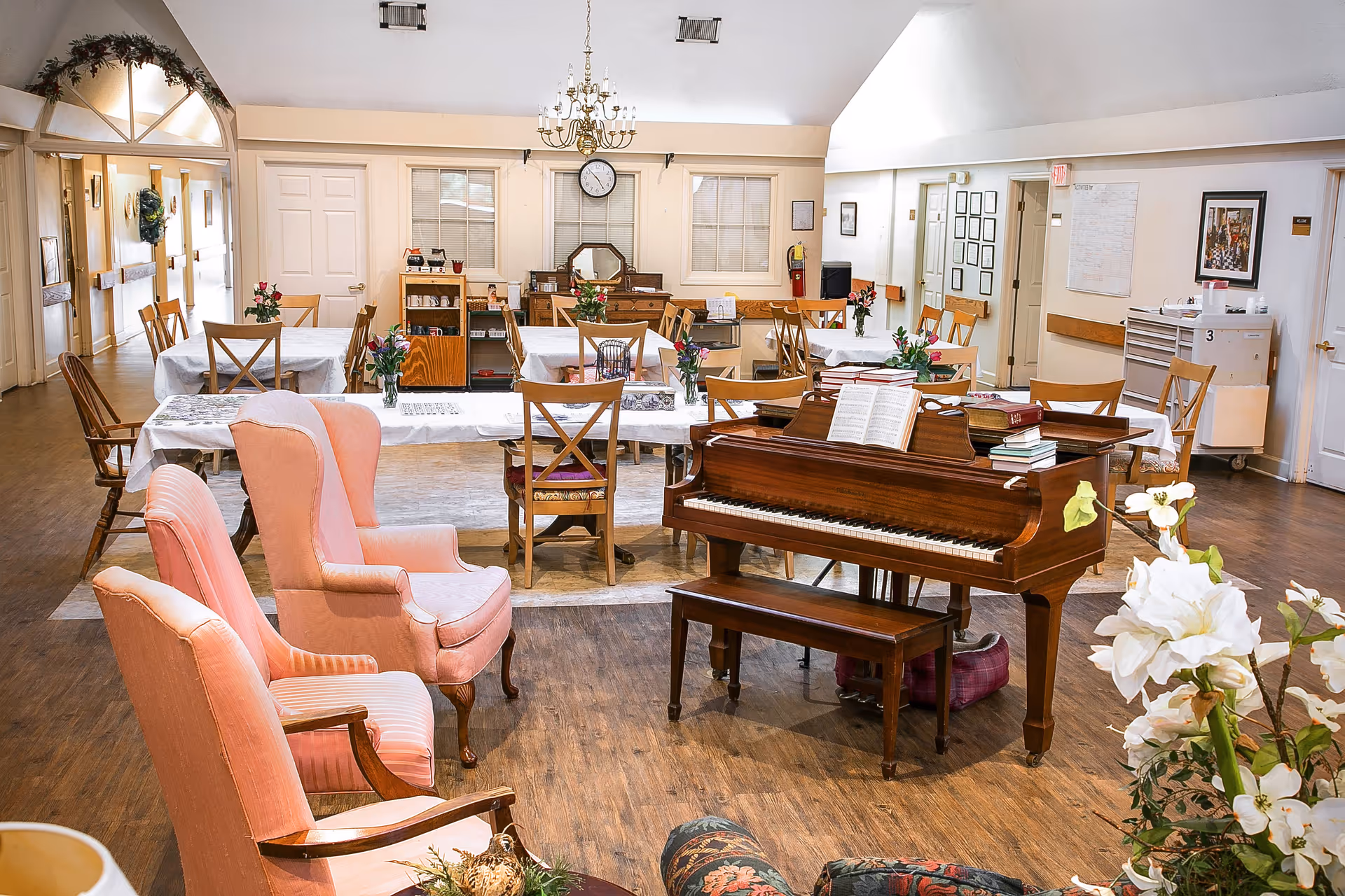 A spacious senior living common area with wooden floors, several tables covered with white tablecloths and decorated with flower vases, a wooden piano with sheet music and books on top, and comfortable upholstered chairs in soft pink tones. The room has a high ceiling with a chandelier, windows with blinds, and various framed pictures on the walls.